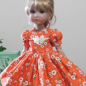 May include: A doll in an orange dress adorned with white floral patterns. The dress features short, puffed sleeves and a white bow at the waist. The doll has blonde hair and is wearing pearl earrings.