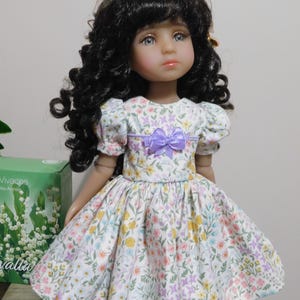 May include: A doll with dark curly hair and blue eyes wearing a floral print dress with a purple bow. The dress features a white background with colorful flowers in shades of pink, yellow, and purple. The doll is posed in a well-lit setting.