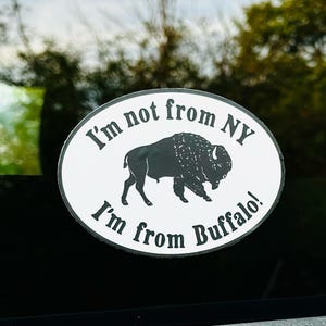May include: Oval white sticker with black text and a black bison silhouette. The text reads "I'm not from NY, I'm from Buffalo!" The sticker is affixed to a dark surface, with a blurred green background.