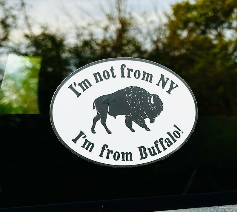 May include: Oval white sticker with black text and a black bison silhouette. The text reads "I'm not from NY, I'm from Buffalo!" The sticker is affixed to a dark surface, with a blurred green background.