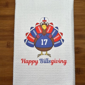 May include: A white waffle-weave kitchen towel featuring a graphic of a turkey wearing a football helmet and a blue jersey with the number 17. The turkey's tail feathers are made of red and blue footballs. The text reads Happy Billsgiving.