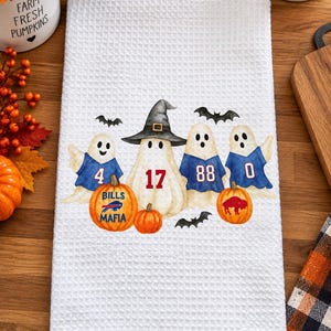 May include: A white waffle-weave kitchen towel featuring a graphic of four ghosts wearing blue football jerseys with the numbers 4, 17, 88, and 0. Two orange pumpkins are included, one with the text Bills Mafia and a buffalo logo.