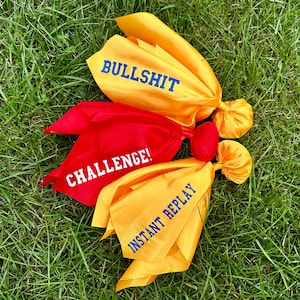 May include: Three yellow fabric pouches with blue embroidered text. The pouches are tied together with a red fabric pouch with white embroidered text. The text on the pouches reads "Bullshit", "Instant Replay", and "Challenge!"