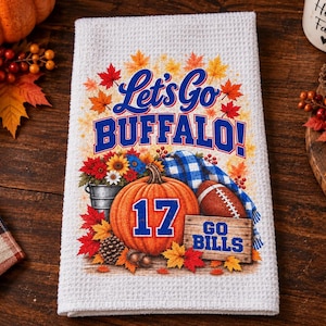May include: A white waffle-weave kitchen towel featuring a colorful autumn-themed graphic with the text Let's Go BUFFALO!, the number 17 on a pumpkin, and a wooden sign reading GO BILLS, surrounded by fall leaves, a football, and flowers.