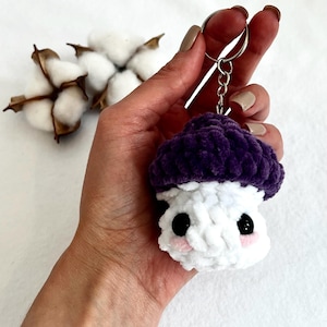 May include: A purple and white crocheted mushroom keychain with black eyes and pink cheeks. The keychain is attached to a silver key ring.