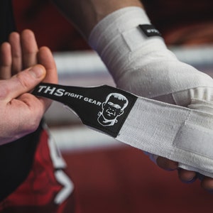 Puede incluir: Vendas blancas para boxeo con una correa negra que dice "THS FIGHT GEAR" y una imagen de dibujos animados de un monstruo.