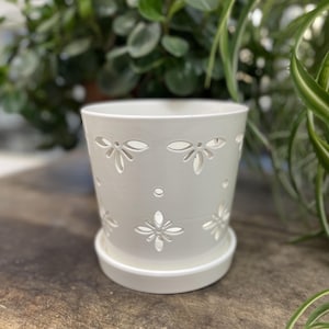 May include: A white ceramic plant pot with a matching saucer, featuring a series of cut-out floral and circular patterns around the exterior. The pot has a smooth, glossy finish and a tapered cylindrical shape, designed to hold small indoor plants.