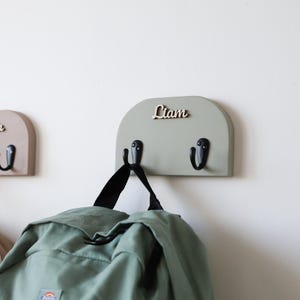 May include: Three wall-mounted arched wooden coat racks in beige, dusty pink, and sage green. Each rack features two black metal hooks and a personalized wooden name cutout reading Amelia and Liam, with one name partially obscured by the angle.