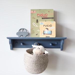 May include: A navy blue wooden shelf with a car-shaped decoration, two books with the word "PUCIO", and a woven basket containing a stuffed animal. The shelf is mounted on a white wall.
