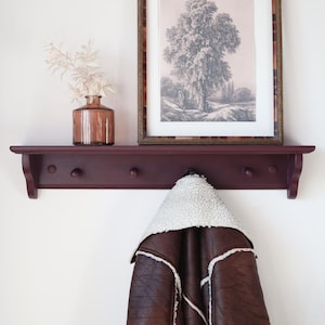 Könnte beinhalten: Ein dunkelburgunderfarbenes Wandregal mit drei Haken, das einen gerahmten Schwarz-Weiß-Baumdruck, eine braune Glasvase mit weißen Stielen und eine braune Lederjacke mit weißem Shearling-Futter trägt. Das Regal ist an einer weißen Wand befestigt.