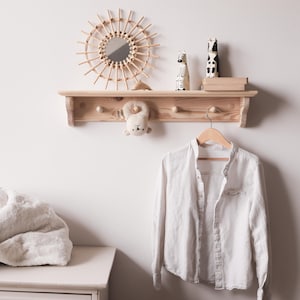 以下が含まれることがあります： サンバーストミラー、装飾的な置物、ぬいぐるみのある明るい色の木製棚。白いストライプのシャツが下にぶら下がっています。白いドレッサーとふわふわの毛布が前景にあります。