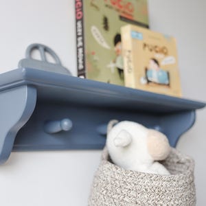 May include: A blue wooden shelf with two pegs, holding books and a toy car. A woven basket with a stuffed animal is below the shelf. The shelf is mounted on a white wall.