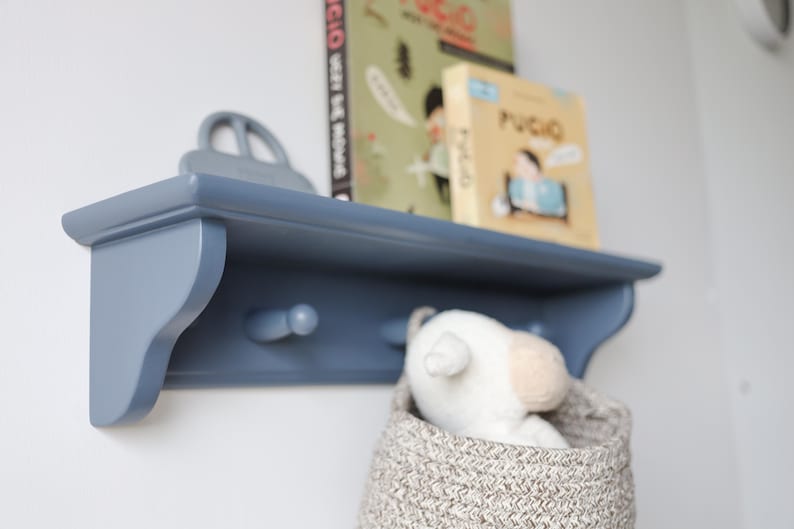 May include: A blue wooden shelf with two pegs, holding books and a toy car. A woven basket with a stuffed animal is below the shelf. The shelf is mounted on a white wall.