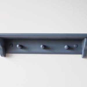 May include: A blue-gray wall shelf with three rounded pegs for hanging items. The shelf has a simple, classic design with a small lip and decorative side supports. The shelf is painted in a matte finish.