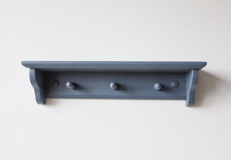 May include: A blue-gray wall shelf with three rounded pegs for hanging items. The shelf has a simple, classic design with a small lip and decorative side supports. The shelf is painted in a matte finish.