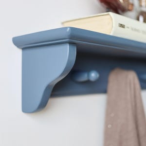 May include: A blue wooden wall shelf with a curved design, featuring two pegs below and a book on top. A tan scarf hangs from the pegs. The shelf is mounted on a white wall.