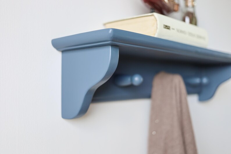 May include: A blue wooden wall shelf with a curved design, featuring two pegs below and a book on top. A tan scarf hangs from the pegs. The shelf is mounted on a white wall.