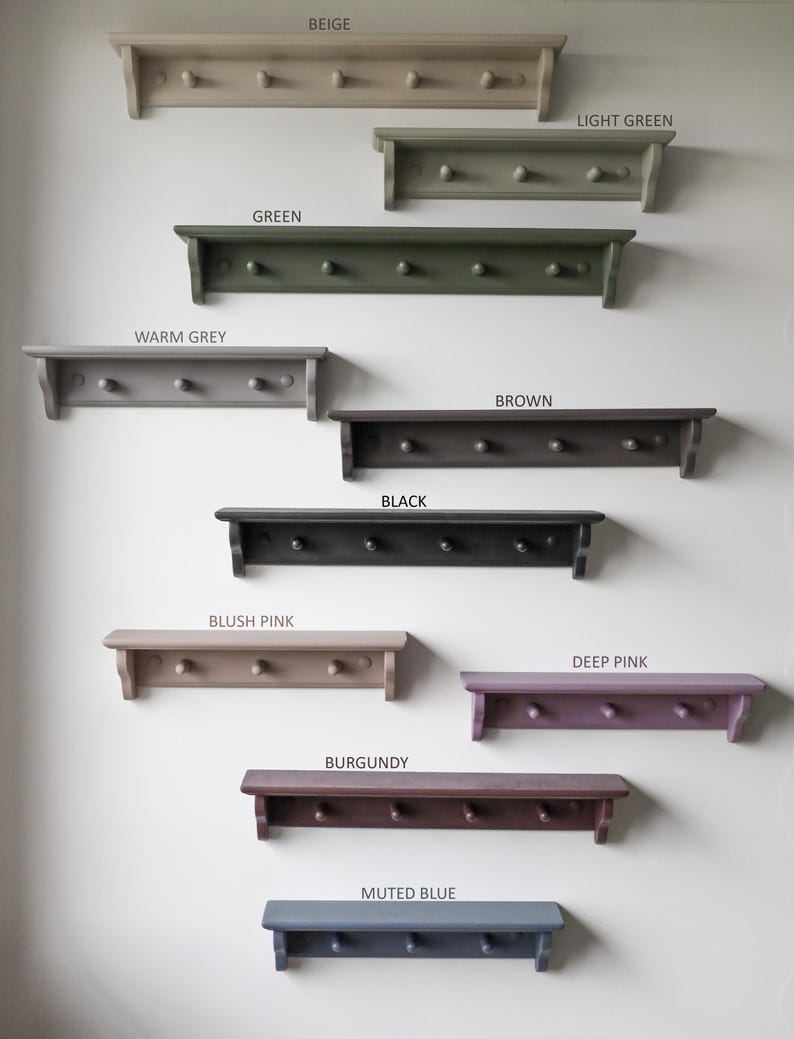 May include: A collection of wooden wall shelves with pegs, each painted in a different color. Colors include beige, light green, green, warm grey, brown, black, blush pink, deep pink, burgundy, and muted blue. Each shelf has a label with its color name.