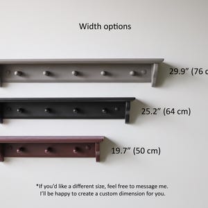 May include: Three wooden wall shelves with pegs in gray, black, and burgundy. The shelves are 29.9" (76 cm), 25.2" (64 cm), and 19.7" (50 cm) wide. Text: Width options. If you'd like a different size, feel free to message me.