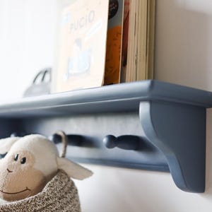 May include: A navy blue wooden shelf with two pegs, holding books and a stuffed animal in a woven basket. The shelf is mounted on a white wall, creating a clean and organized storage solution.