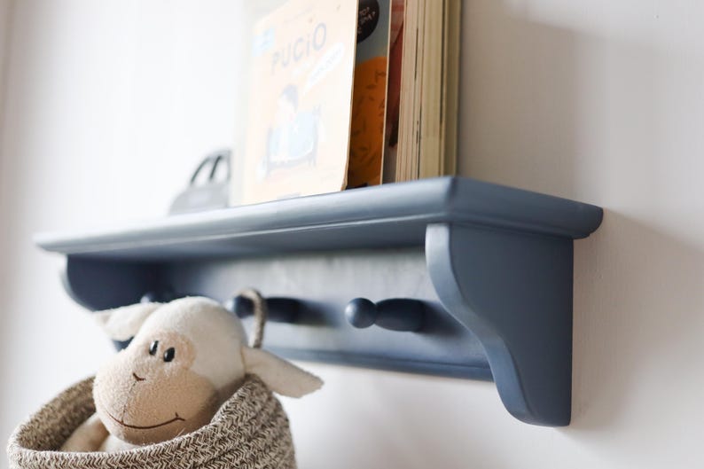 May include: A navy blue wooden shelf with two pegs, holding books and a stuffed animal in a woven basket. The shelf is mounted on a white wall, creating a clean and organized storage solution.