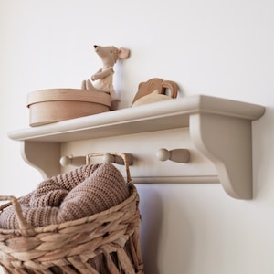 Könnte beinhalten: Ein beiges Wandregal mit einem Weidenkorb und einer runden Box. Ein Stoffmaus-Spielzeug und ein Holzspielzeug befinden sich auf dem Regal. Der Korb ist mit einer braunen Strickdecke gefüllt. Das Regal hat zwei Holzstifte.