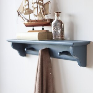 May include: A blue wall shelf with two hooks, holding a brown towel. A decorative wooden sailboat, a book, and a glass bottle with contents are displayed on the shelf. The shelf is mounted on a white wall.