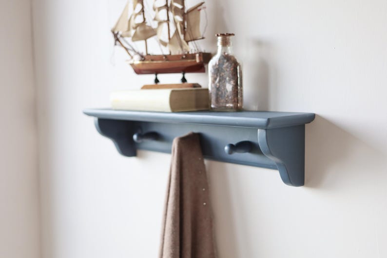 May include: A blue wall shelf with two hooks, holding a brown towel. A decorative wooden sailboat, a book, and a glass bottle with contents are displayed on the shelf. The shelf is mounted on a white wall.