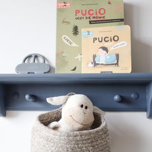 May include: A navy blue wooden shelf with three pegs, holding two children's books, a gray car-shaped toy, and a woven basket with a stuffed lamb. The books have Polish text and illustrations.