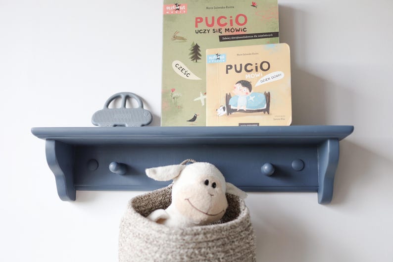 May include: A navy blue wooden shelf with three pegs, holding two children's books, a gray car-shaped toy, and a woven basket with a stuffed lamb. The books have Polish text and illustrations.