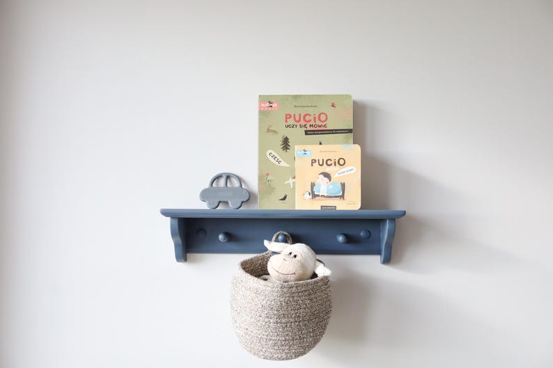 May include: A navy blue wooden shelf with a car-shaped decoration, two books with the word "PUCIO", and a woven basket containing a stuffed animal. The shelf is mounted on a white wall.