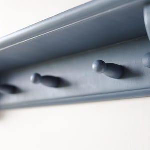 May include: A blue-gray wooden shelf with three peg hooks. The shelf has a simple design with a flat top and a slightly curved front edge. The peg hooks are evenly spaced and are a similar color to the shelf.