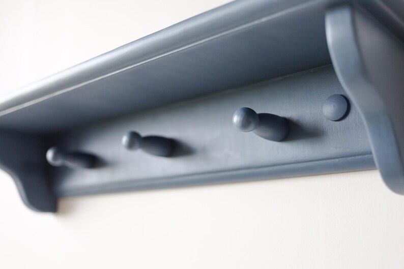 May include: A blue-gray wooden shelf with three peg hooks. The shelf has a simple design with a flat top and a slightly curved front edge. The peg hooks are evenly spaced and are a similar color to the shelf.