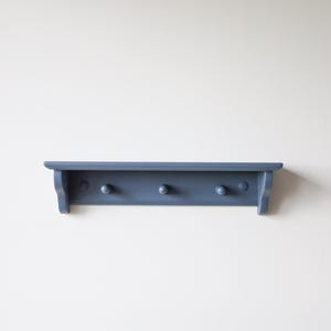 May include: A blue-gray wooden shelf with three pegs. The shelf has a simple design with a flat top and a slightly curved front edge. The pegs are evenly spaced along the bottom of the shelf.