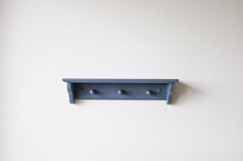 May include: A blue-gray wooden shelf with three pegs. The shelf has a simple design with a flat top and a slightly curved front edge. The pegs are evenly spaced along the bottom of the shelf.
