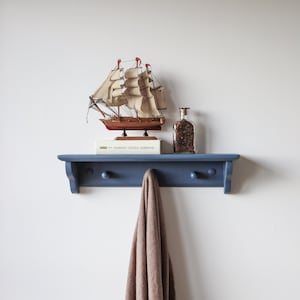 May include: A navy blue wall shelf with two wooden knobs, holding a model sailboat, a glass bottle, and a book. A brown towel hangs from the shelf. The shelf is mounted on a white wall.