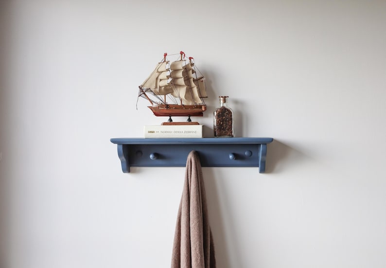 May include: A navy blue wall shelf with two wooden knobs, holding a model sailboat, a glass bottle, and a book. A brown towel hangs from the shelf. The shelf is mounted on a white wall.