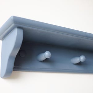 May include: A blue-gray wooden wall shelf with three peg hooks. The shelf has a decorative edge and is mounted on a white wall. The pegs are rounded and evenly spaced, providing a functional and stylish storage solution.