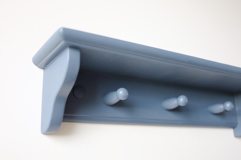 May include: A blue-gray wooden wall shelf with three peg hooks. The shelf has a decorative edge and is mounted on a white wall. The pegs are rounded and evenly spaced, providing a functional and stylish storage solution.