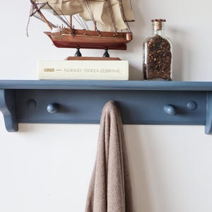 May include: A blue wall shelf with three wooden pegs, holding a small model sailboat, a book, and a glass jar filled with dark objects. A tan scarf hangs from one of the pegs. The shelf is mounted on a white wall.