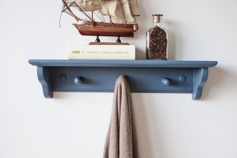 May include: A blue wall shelf with three wooden pegs, holding a small model sailboat, a book, and a glass jar filled with dark objects. A tan scarf hangs from one of the pegs. The shelf is mounted on a white wall.