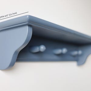 May include: A blue painted wooden shelf with three pegs below. The shelf is 4.6 inches (11.7cm) deep and 4.6 inches (11.7cm) high. The shelf is mounted on a white wall.