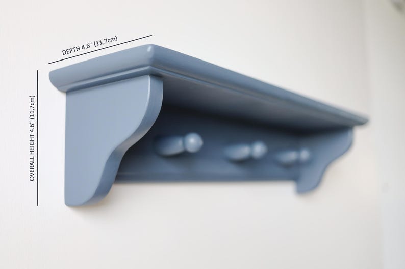 May include: A blue painted wooden shelf with three pegs below. The shelf is 4.6 inches (11.7cm) deep and 4.6 inches (11.7cm) high. The shelf is mounted on a white wall.