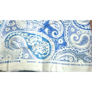 May include: A piece of fabric featuring a blue and white paisley pattern with floral accents. The bottom edge includes the printed text: "AMASURA" AN EXCLUSIVE DESIGN BY RICHARD SMITH FOR JIM THOMPSON, along with a series of small blue circles.