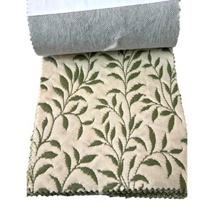 May include: A fabric sample with a cream-colored background and a repeating pattern of green leaves and vines. The reverse side of the fabric is a solid gray. The edges of the fabric are unfinished.