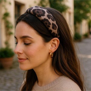May include: A padded headband with a brown and black leopard print pattern. The headband is worn on the head. The person is also wearing gold hoop earrings.