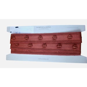 May include: A spool of rust-colored decorative trim featuring a repeating pattern of small, raised circular floral motifs. The trim is wrapped around a white cardboard card labeled with the text Samuel & Sons Passementerie at the top.