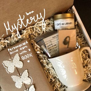 May include: A brown box filled with a mystery gift box for a book lover. The box contains a wrapped book with a book-page butterfly decoration, a white mug, a candle that says "Cafe au Library", and a book-shaped key chain.