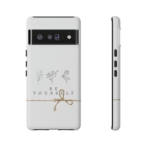 May include: White phone case with a black camera lens and a brown twine bow. The case has a minimalist design with the text "BE YOURSELF" and three small floral illustrations.