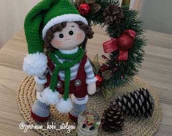 Puede incluir: Una muñeca de elfo de crochet con un gorro verde, una camisa a rayas rojas y blancas y una bufanda verde. La muñeca está de pie sobre una estera tejida con una corona de Navidad y piñas.